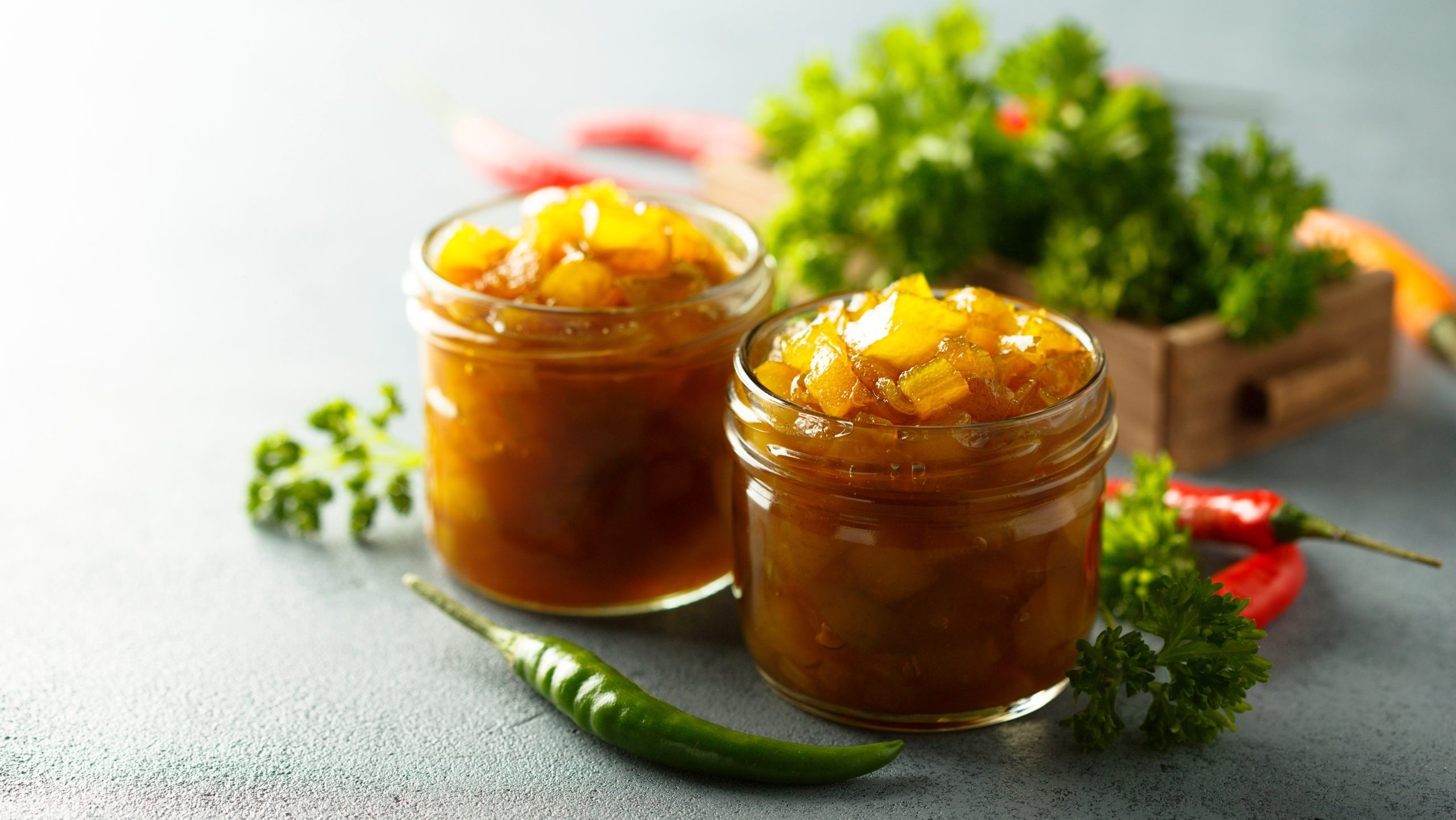 Zwei Gläser Apfelchutney mit gelber Paprika in Frontalaufnahme. Eine grüne Chili davor. Krause Petersilie um die Gläser drapiert.