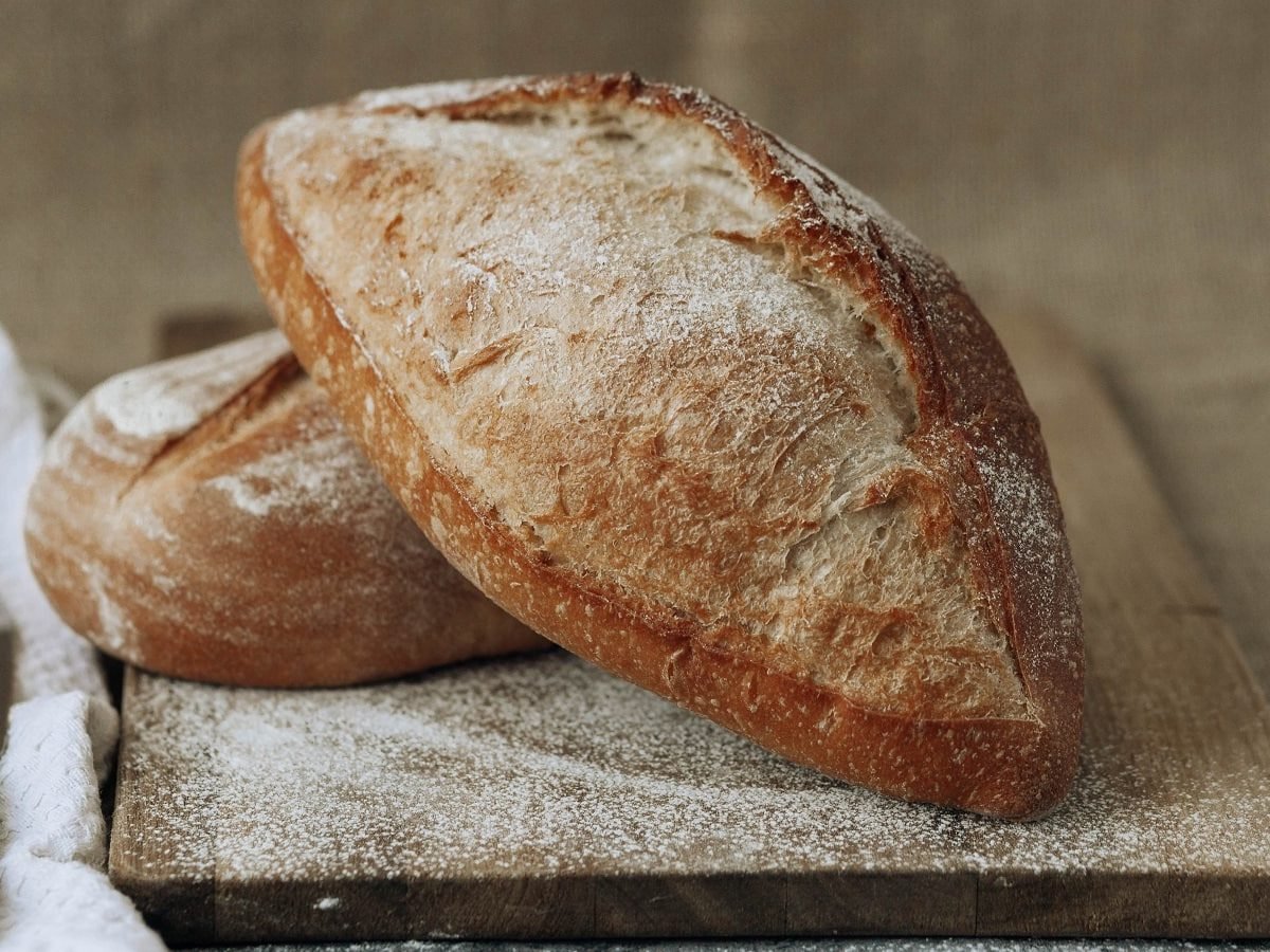 Zwei Baguette-Brötchen mit Kräuter der Provence auf einem Holzschneidebrett neben einem Brotmesser und einem weißen Tuch, vor einem braun-beigenen Hintergrund.