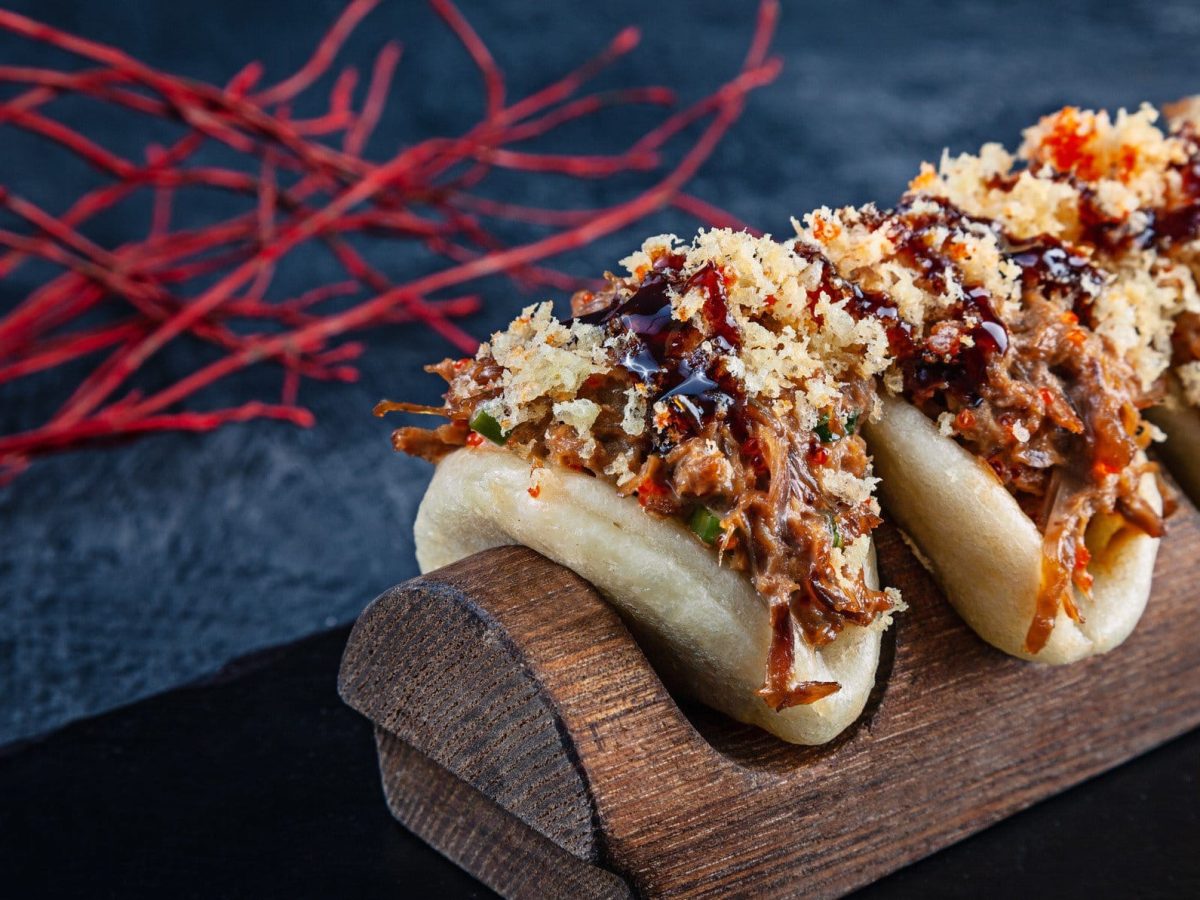 Vier Baos mit Char siu, Crunch und Hoisinsoße in Bambushalterung aus der Frontalsicht. Rote Chilifäden auf blauer Platte im Hintergrund.