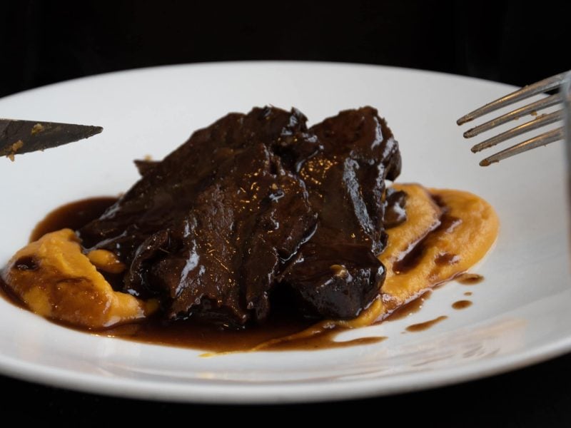 Braten mit Glühweinsoße auf gelbem Püree auf weißem Teller. Im Vordergrund ein Glas halb voll mit Bier. Messer und Gabel am Bildrand. Aufnahme aus der Frontalansicht.