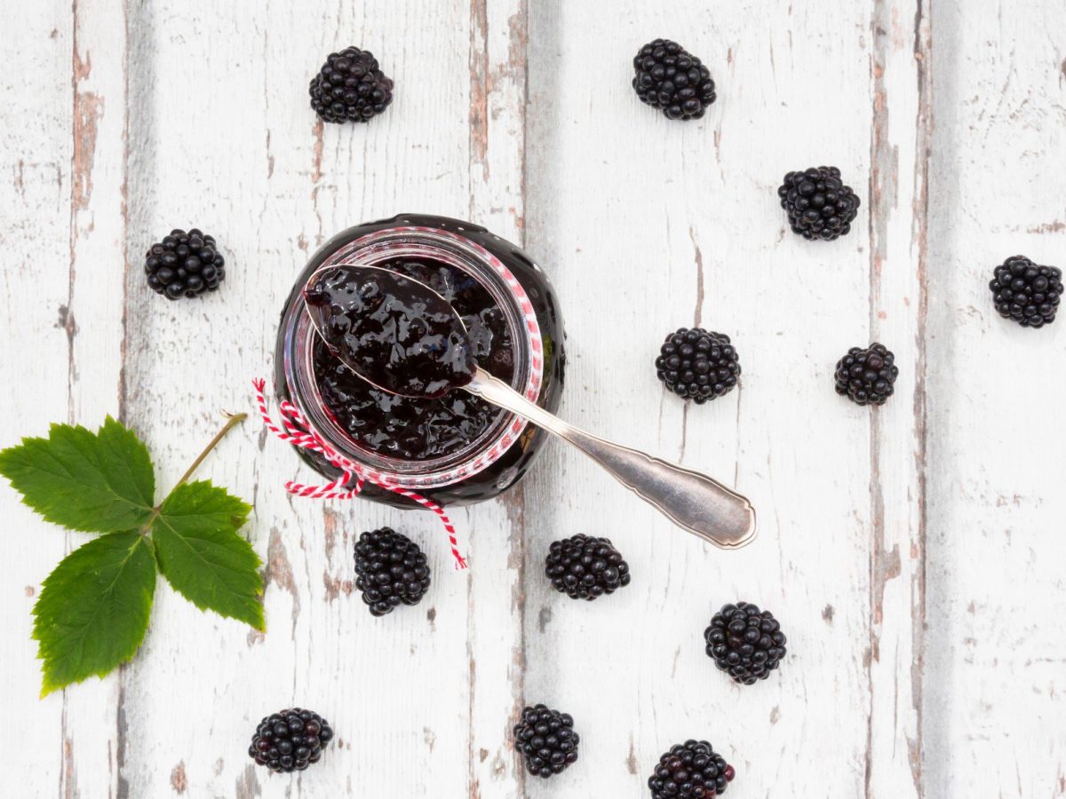 In der Draufsicht sieht man ein Glas Brombeermarmelade mit Chia auf hellem Holz, daneben liegen ein paar Brombeeren und Minze.