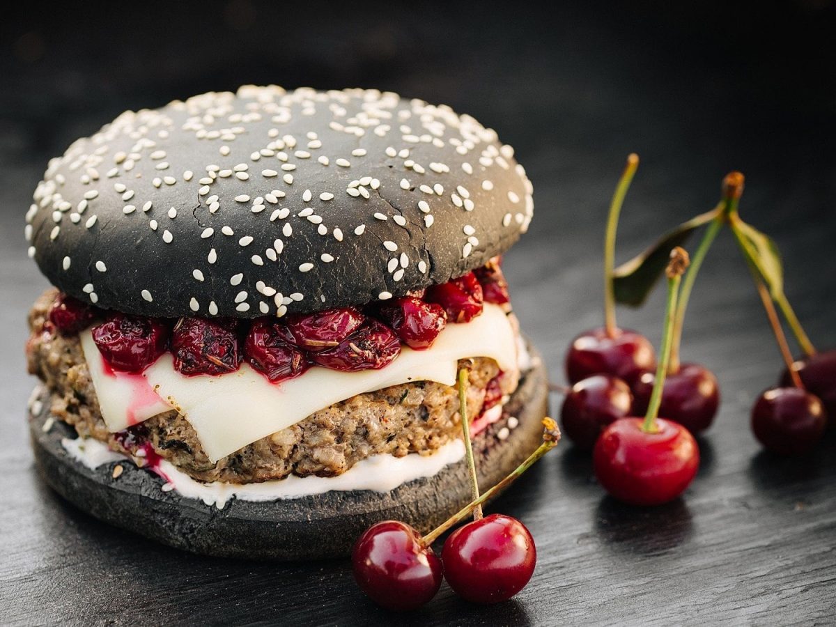 Ein Burger mit Sauerkirschen steht auf einem dunklen Holztisch. Der Burger besteht aus schwarzen Burgerbuns, Rindfleischpatties, Käse und Sauerkirschen.