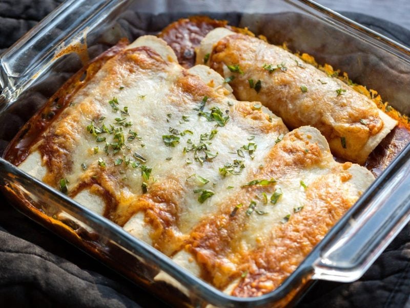 Chili-Käse-Enchiladas in Auflaufform aus Glas auf grauem Topfhandschuh und dunklem Holztisch. Aufnahme aus der Draufsicht.