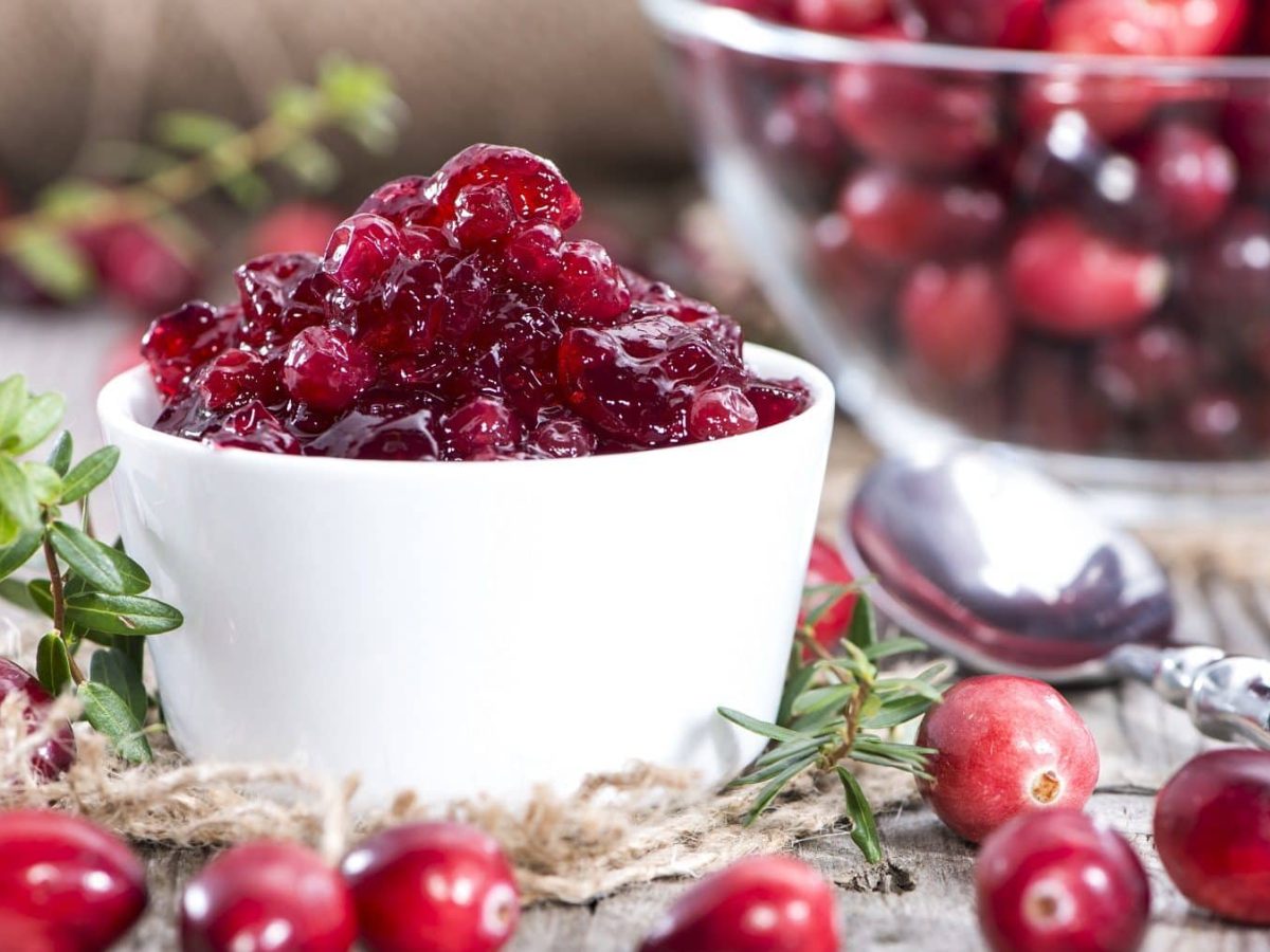 Auf einem Holztisch steht eine Cranberry-Marmelade. Die ist in einem weißen Schälchen. Drumherum liegen Cranberry. Auch eine GLasschüssel mit Cranberrys ist zu sehen.