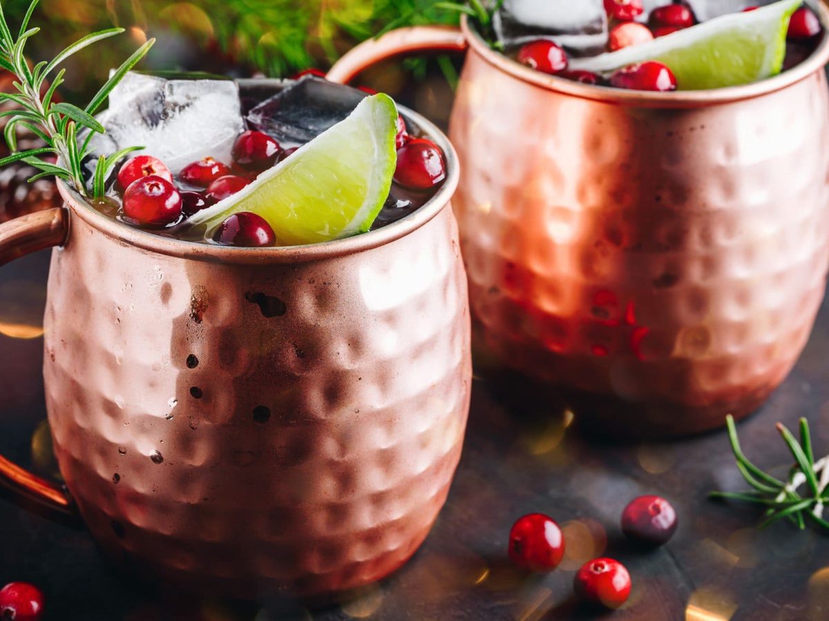 Zwei Kupferbecher mit Cranberry Moscow Mule, Eiswürfeln, Rosmarin und einer Limettenspalte.