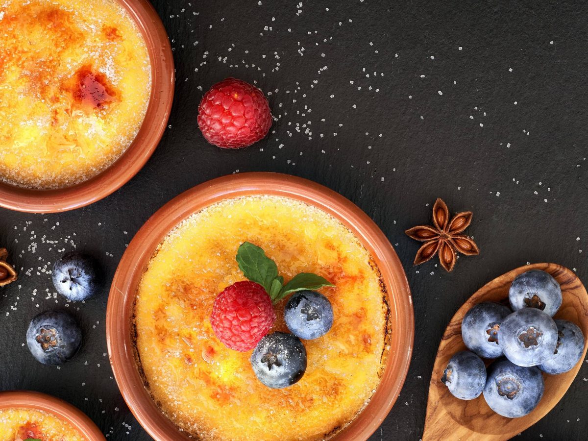 Crema Catalana in Tonförmchen auf schwarzer Platte. Dekoriert mit Blaubeeren, Himbeeren, Minze und Sternanis. Aufnahme aus der Vogelperspektive