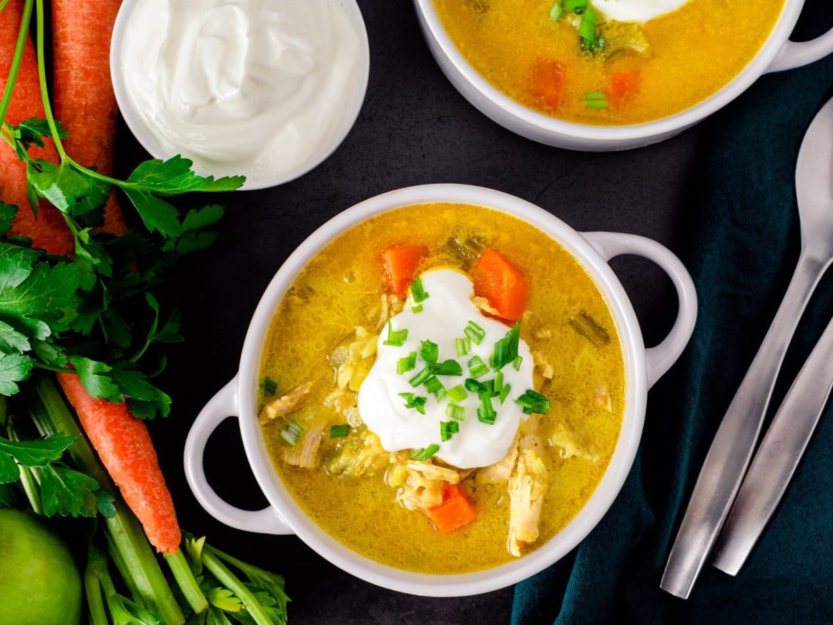 Zwei Mulligatawny Suppe aus Dinner for One in weißer Suppenschale mit zwei Henkeln auf schwarzer Schieferplatte. Auf der Suppe ein Kleks Creme Fraiche und Schnittlauch. Rechts unten zwei Suppenlöffel. Links Karotten, Petersilie und Apfel. Darüber eine kleine weiße Schale mit Creme Fraiche. Aufnahme aus der Vogelperspektive.