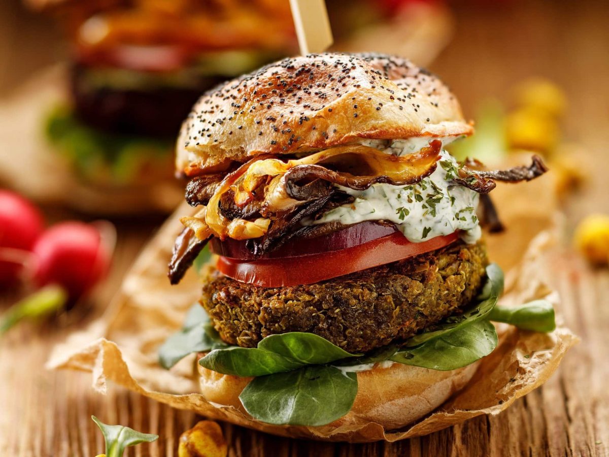 Erbsen-Burger in einem Sesambrötchen mit Tomate, Salat, frischer Remoulade, Röstzwiebeln und veganem Bacon auf einer hölzernen, rustikalen Unterlage