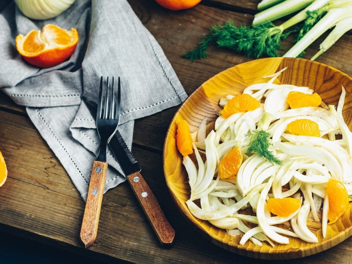 Auf einem Holzteller ist eine Fenchel-Orangen-Salat mit Roquefort-Vinaigrette angerichtet. Neben dem Teller liegen Orangenstücke, ein Messer sowie eine Fenchelknolle.