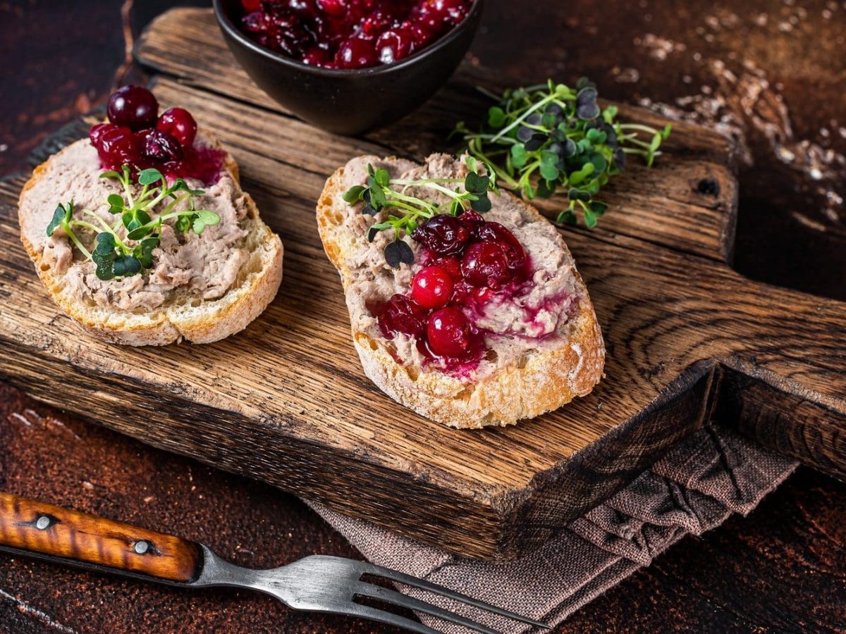 Auf einem Brettchen liegen zwei Scheiben Weißbrot. Diese sind mit Gänse-Rillettes bestrichen. Dazu gibt es Kirschen und Sprossen