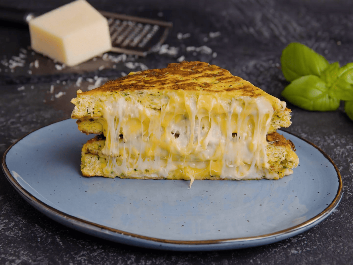 Auf einem blauen Teller liegt halbiertes ein Grilled Cheese mit Zucchinipuffern, dahinter einige Basilikumblätter und Parmesan.