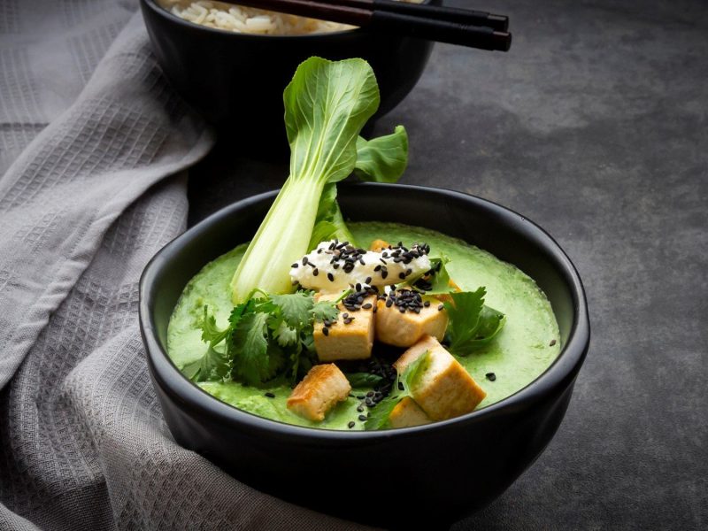 Eine schwarze Schüssel grünes Thai-Curry mit Pak Choi und Tofu steht auf einem dunkelgrauen Untergrund. Im Hintergrund steht eine Schüssel mit Reis und zwei Essstäbchen. Links neben dem Curry liegt ein graues Geschirrtuch.