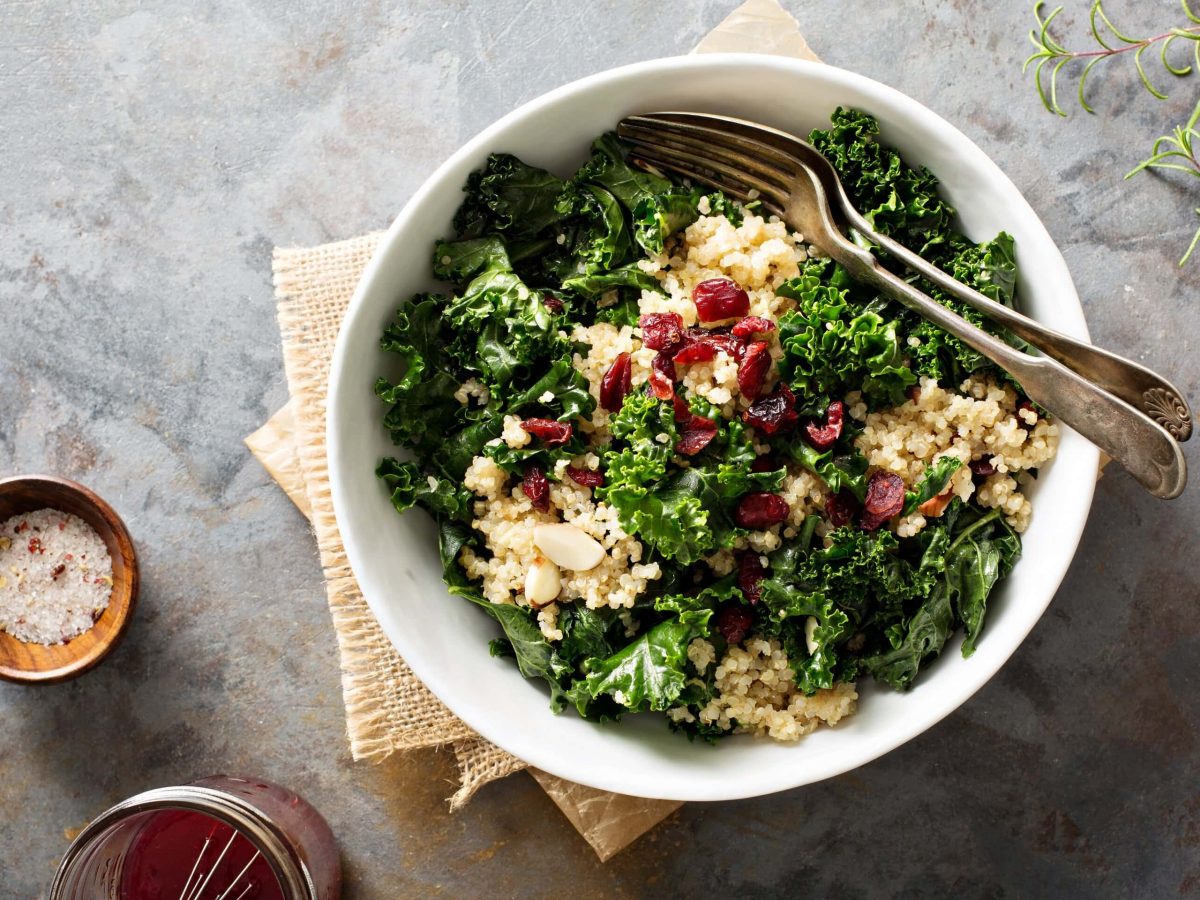 Eine weiße Schüssel Grünkohlsalat mit Quinoa und Cranberrys, darin eine Gabel und ein Löffel, in der Draufsicht. Daneben ein Schälchen Salz und ein Glas Dressing.