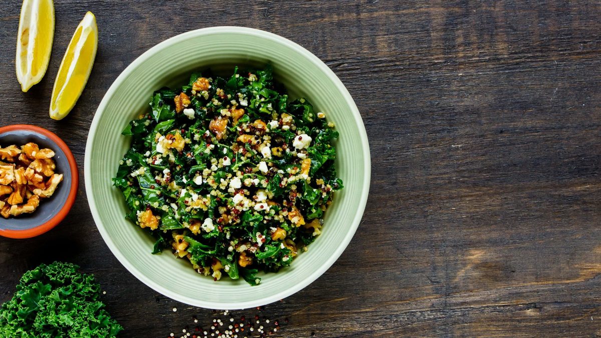 Ein tiefer Teller Grünkohlsalat mit Walnüssen und veganem Feta in der Draufsicht, daneben liegen zwei Zitronenspalten und Grünkohl, außerdem ist eine Schale Walnüsse zu sehen.