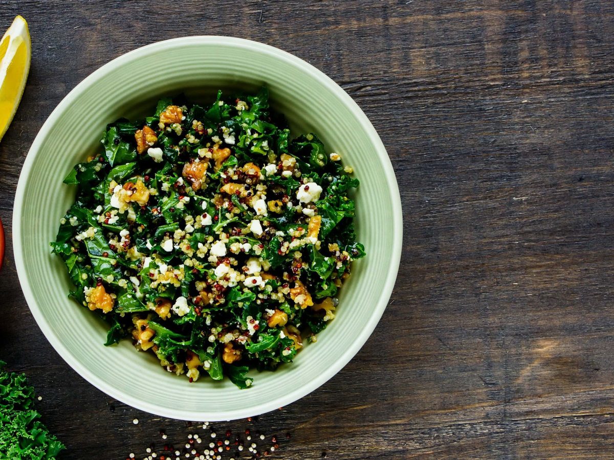 Ein tiefer Teller Grünkohlsalat mit Walnüssen und veganem Feta in der Draufsicht, daneben liegen zwei Zitronenspalten und Grünkohl, außerdem ist eine Schale Walnüsse zu sehen.