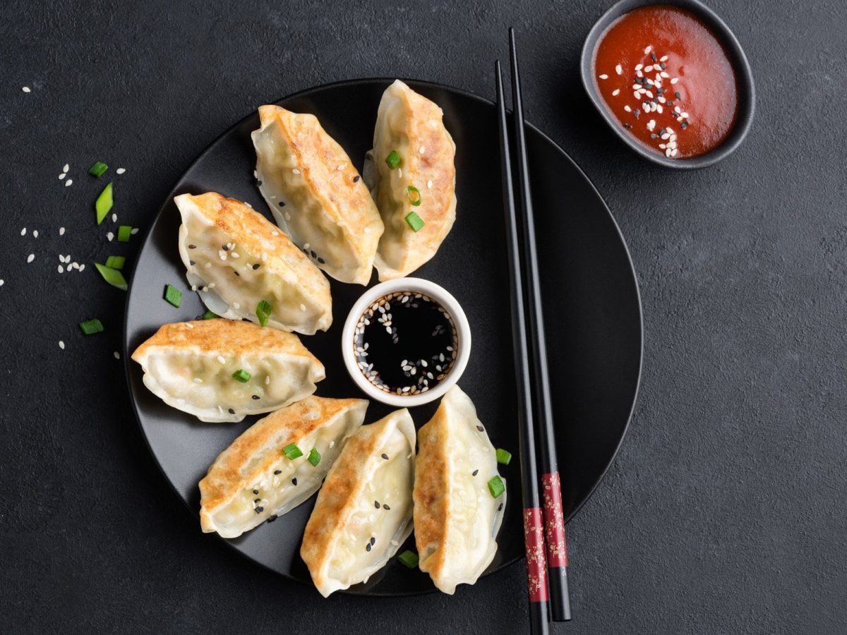 Gyoza mit Hähnchenfüllung mit schwarzen Essstäbchen auf schwarzem Teller und schwarzem Grund. In der Tellermitte eine weiße Schale mit dunklaer Sauce. Rechts oben schwarze Schale mit roter Sauce. Aufnahme aus der Vogelperspektive.