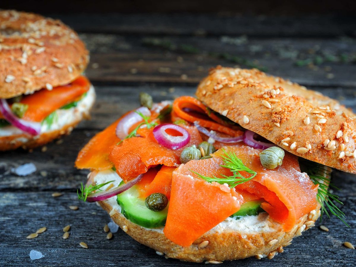 Veganer Karotten-Lachs mit Räuchersalz, Kapern und Öl auf einem Bagel mit Frischkäse, Gurke, Zwiebeln auf einem schwarzen Holzbrett