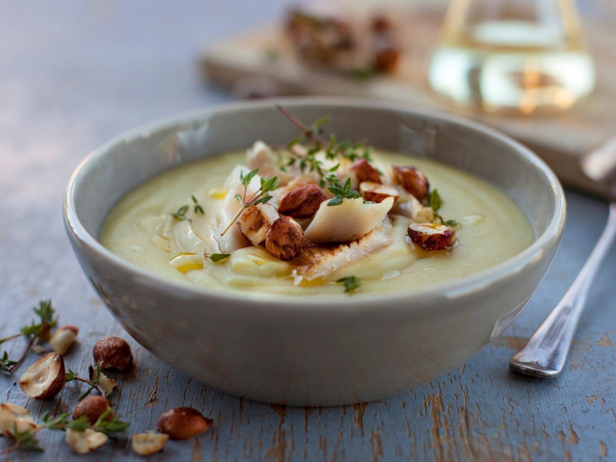 Eine graue Schüssel Kartoffelsuppe mit Räucherfisch und Haselnüssen auf blauem Holz, daneben liegt ein Löffel.