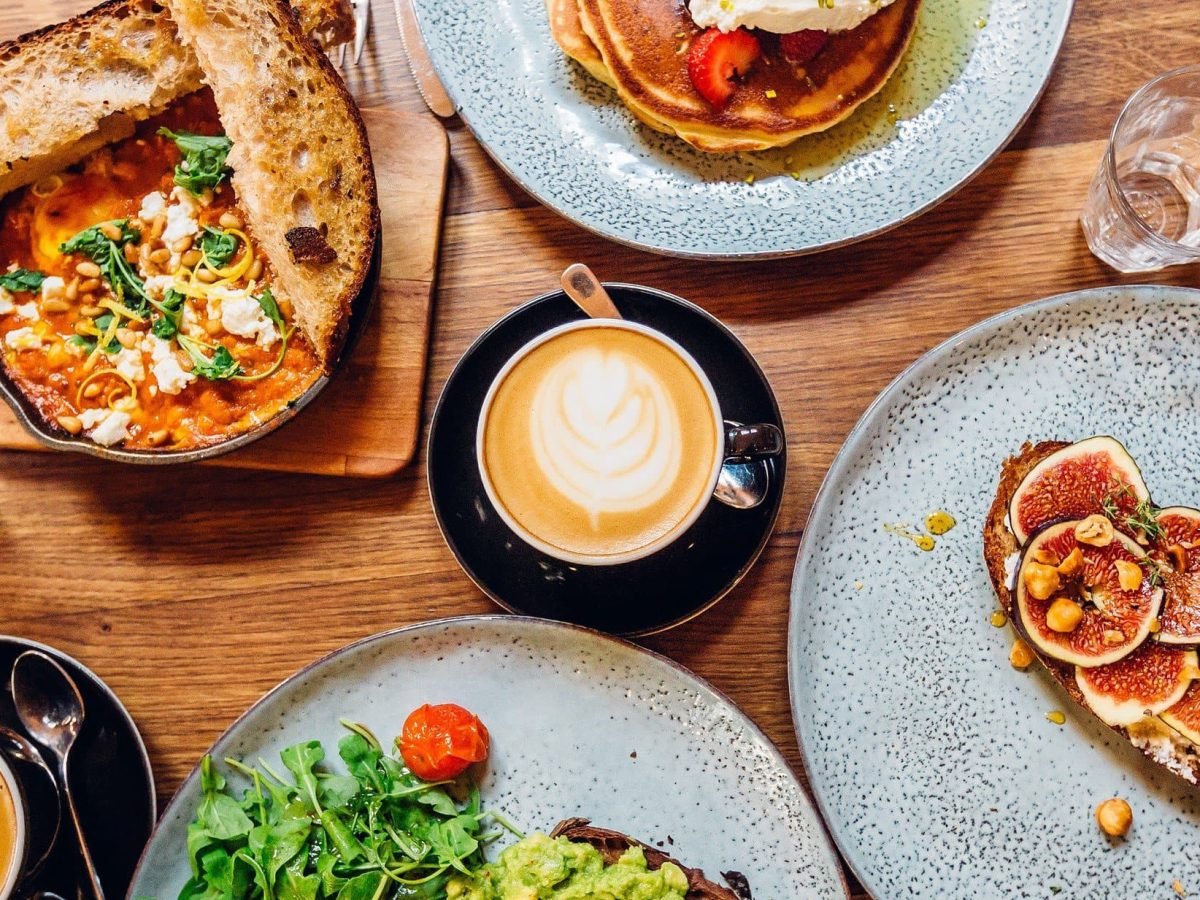 Holztisch auf dem vier Teller serviert sind, ein Teller mit Feigenfrischkäsebrot, ein Teller mit Avocadolachsbrot und Rucolasalat, ein Teller mit selbstgemachten Pancakes und Ahornsoße und ein Teller mit würziger Shakshuka und getoastetem Brot