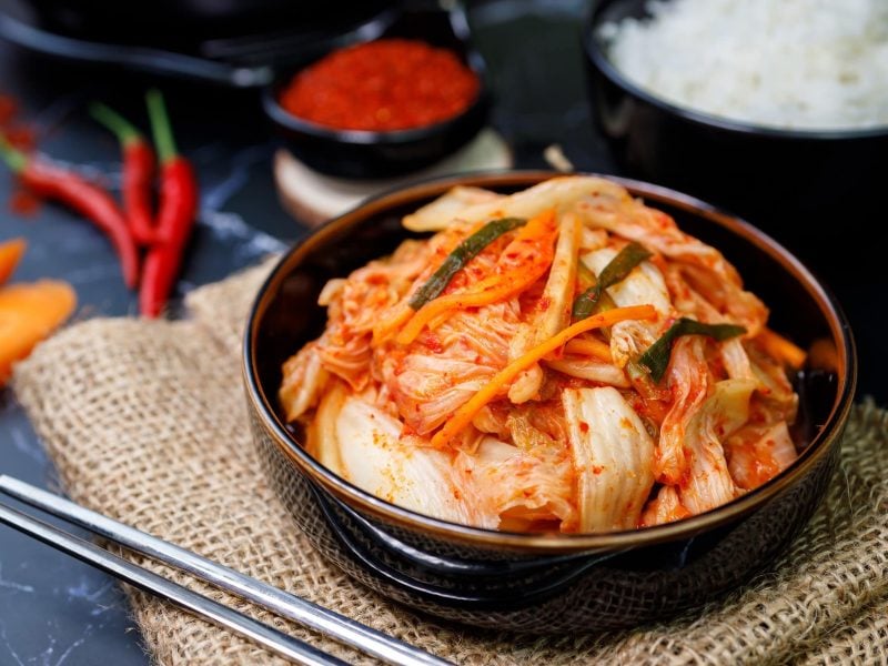 Eingelegter koreanischer Rettich und Kohl, auch Kimchi genannt, auf Reis in einer schwarzen Tonschale, gelegt auf ein Bastnetz mit Reis, Chilli, Karotte und Paprikapulver im Hintergrund.