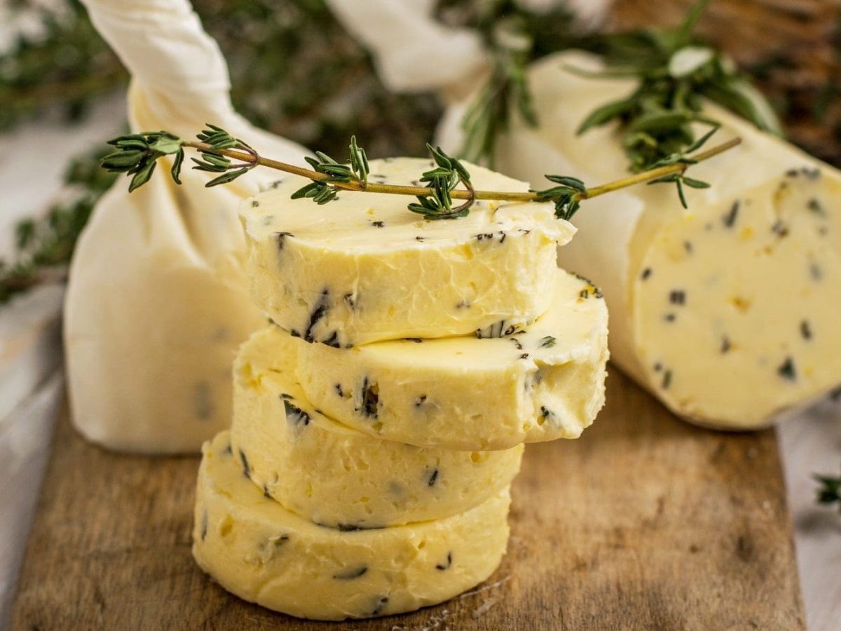 Auf einem Holzbrett liegt eine Kräuterbutter mit Limette. Die Butter ist in Pergamentpaier gerollt und mit Tyhmianzweigen dekoriert.