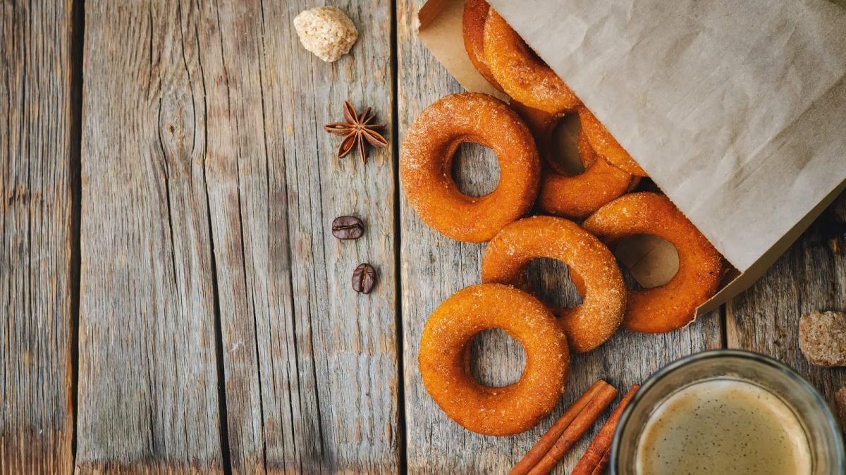 Eine Tüte aus der Kürbis-Donuts fallen, die mit Zimt-Zucker bestreut sind, neben Gewürzen und einer Tasse Kaffe, vor einem Holzhintergrund, von oben.