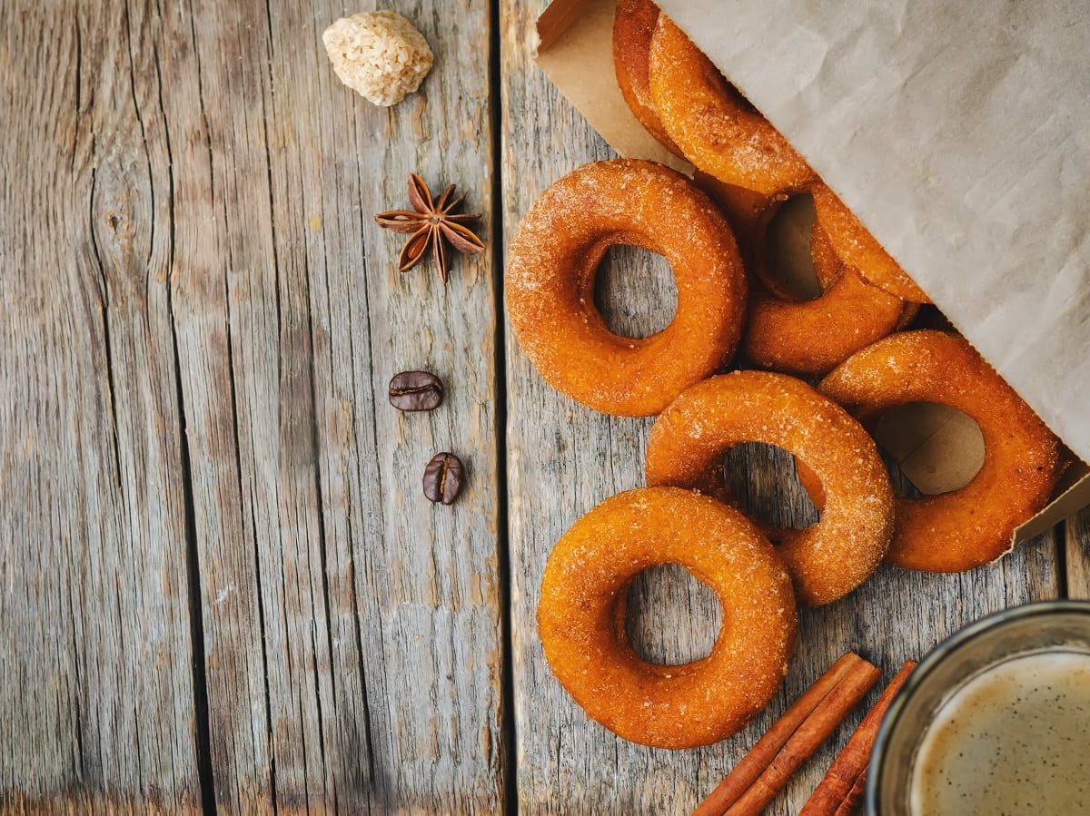 Eine Tüte aus der Kürbis-Donuts fallen, die mit Zimt-Zucker bestreut sind, neben Gewürzen und einer Tasse Kaffe, vor einem Holzhintergrund, von oben.