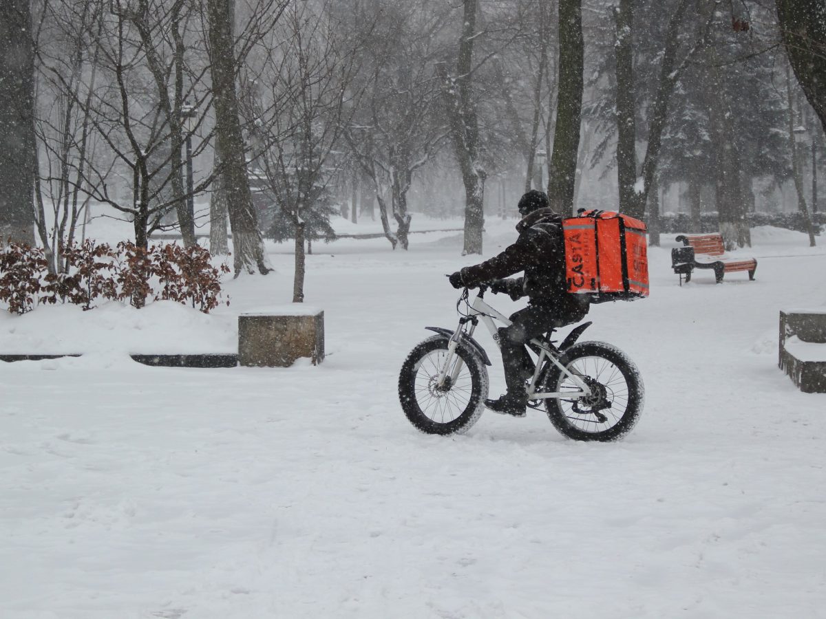 Lieferant mit Rucksack auf Fahhrad fährt durch einen schneebedeckten Park bei Schneefall. Aufnahme aus der Ferne.