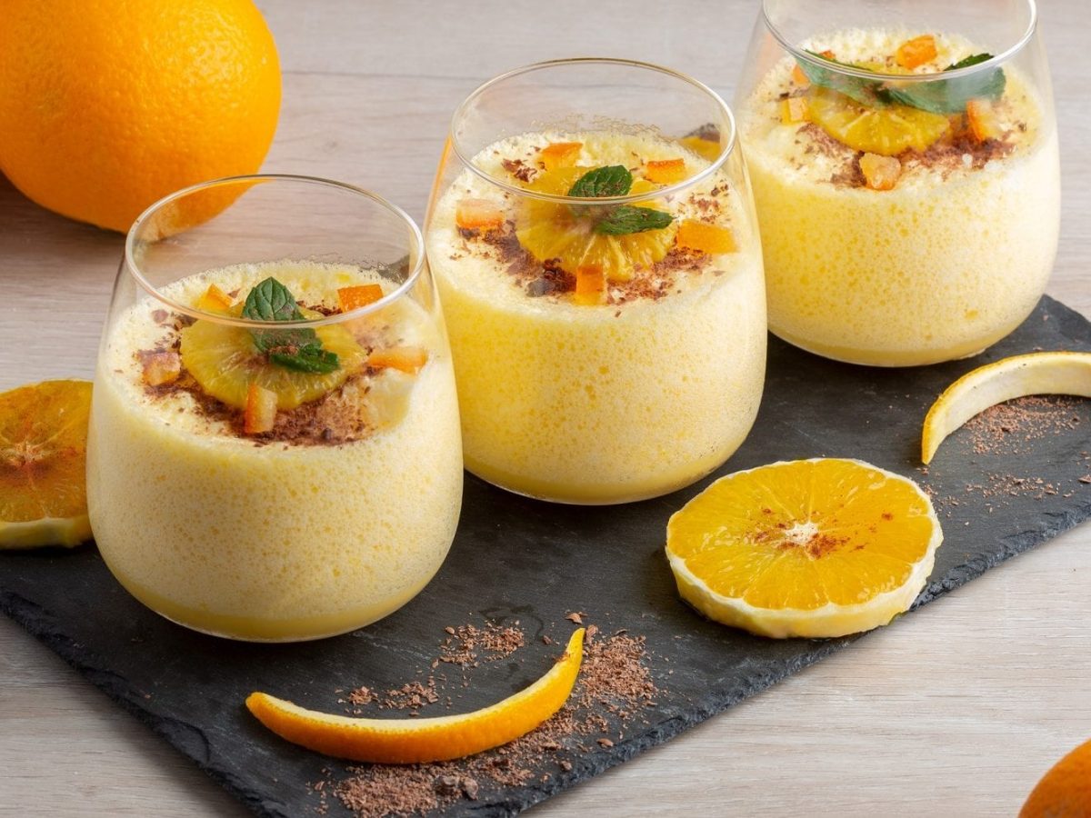 Auf einer Schiefertafel stehen drei Gläser mit Orangen-Mousse. Daneben liegen Orangen-Scheiben sowie drei Orangen.