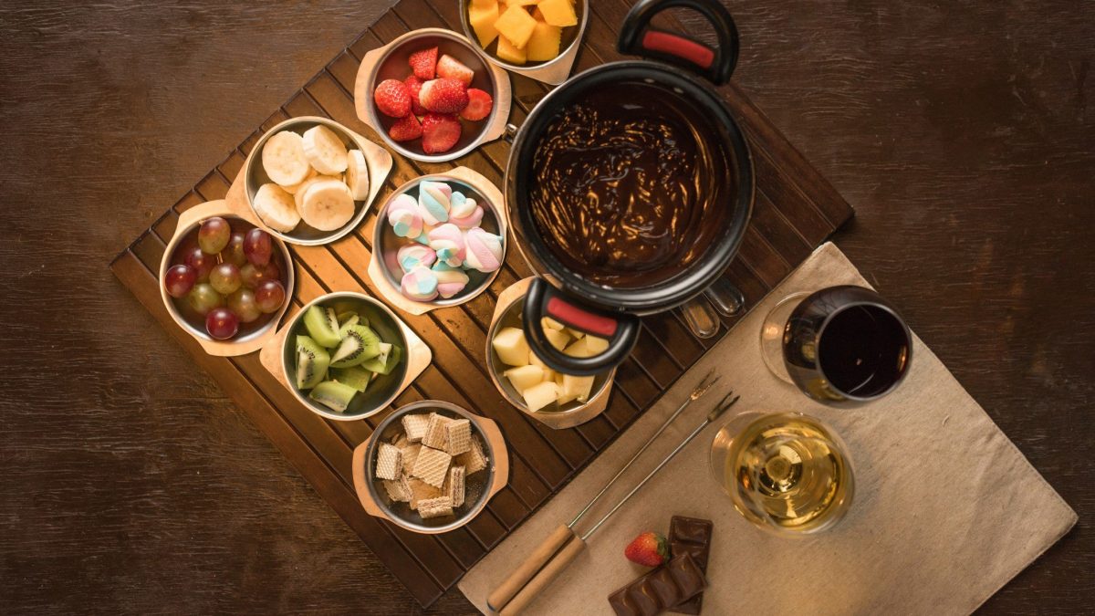 Mon Chéri Fondue Schokoladen Fondue mit diversen Früchten und bunten Marchmellows in Schalen auf Holzbrett auf Holttafel. Daneben je ein Glas Rotwein und Weißwein sowie Fonduegabeln auf hellem Holzbrett. Aufnahme aus der Vogelperspektive.