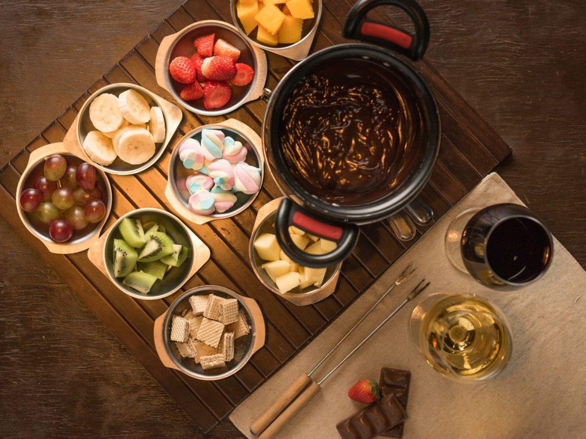 Mon Chéri Fondue Schokoladen Fondue mit diversen Früchten und bunten Marchmellows in Schalen auf Holzbrett auf Holttafel. Daneben je ein Glas Rotwein und Weißwein sowie Fonduegabeln auf hellem Holzbrett. Aufnahme aus der Vogelperspektive.