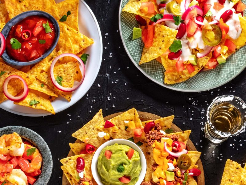 Viele Teller gefüllt mit Nachos, dem Tex-Mex-Gericht aus Tortilla-Chips, verschiedenen Zutaten und überbackenem Käse, neben Mais, Limetten, Salz, Schalne mit Soßen und einem Glas Tequila vor einem dunklen Hintergrund, von oben.