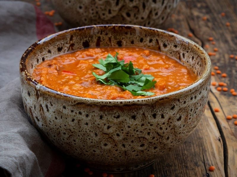 Eine Schüssel Ofengemüse-Suppe bestreut mit frischer Petersilie oder Koriander auf einem Holztisch, daneben ein Geschirrtuch.