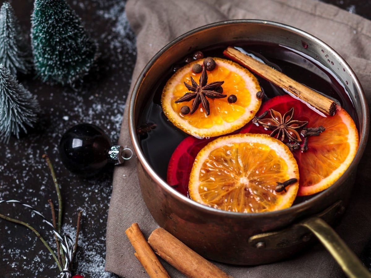 Selbst gemachtes Glühweingewürz: Ein Topf mit Glühwein, Orangen, Zimt und Anis steht auf dunklem Untergrund, daneben kleine Deko-Bäume und Kugeln.