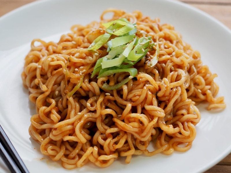 TikTok-Ramen mit Lauchzwiebel angerichtet auf einem weißen Teller. Der Teller steht auf einem Holztisch. Neben den Nudeln liegen zwei schwarze Esstäbchen.