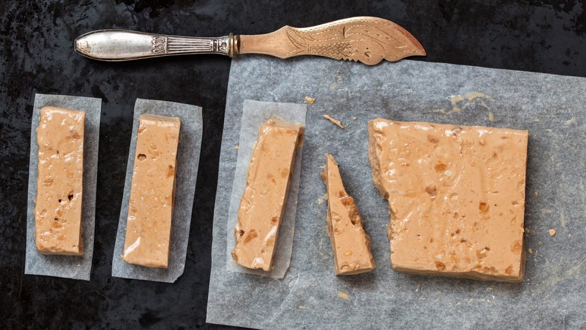 Ein in Stücke geschnittenes Turrón de Navidad neben einem Messer, vor einem dunklen Hintergrund, von oben.