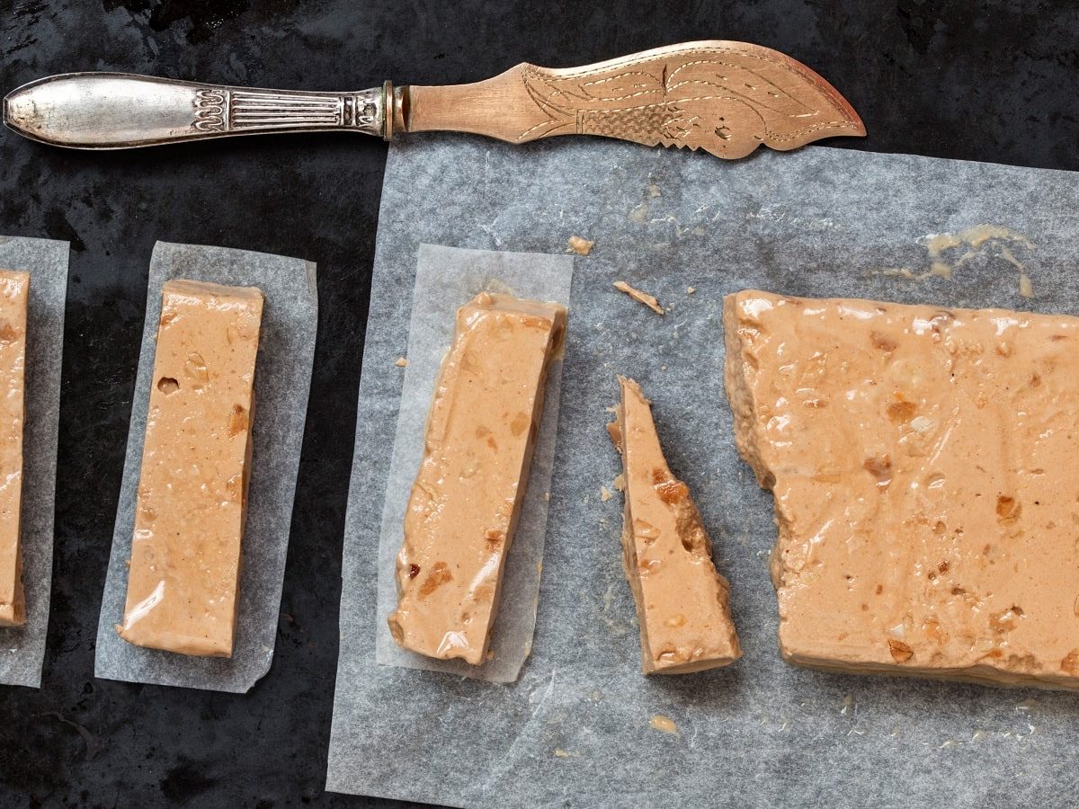 Ein in Stücke geschnittenes Turrón de Navidad neben einem Messer, vor einem dunklen Hintergrund, von oben.