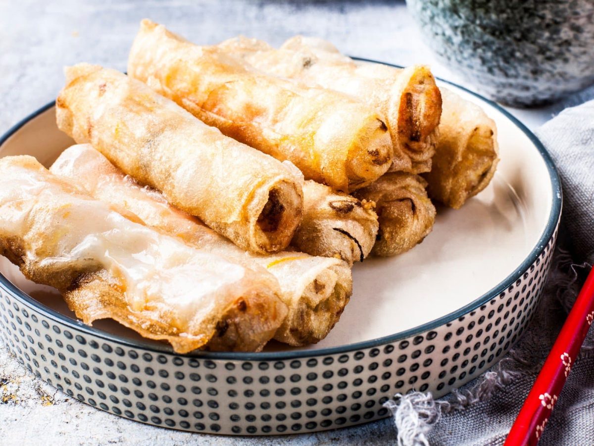 In einer kleinen Schüssel liegen einige vegane Frühlingsrollen, daneben rote Essstäbchen.