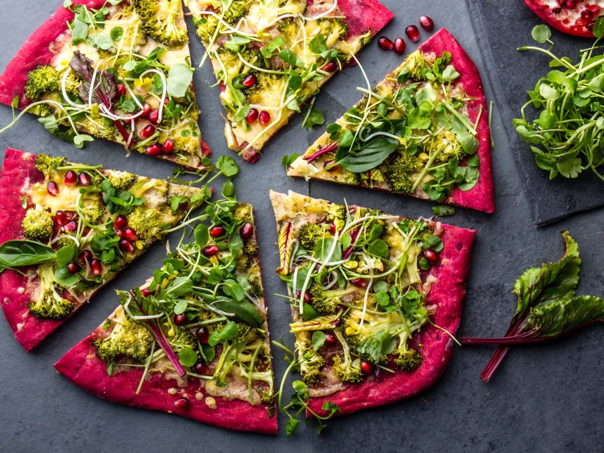 Eine geschnittene Rotkohl-Pizza mit üppigem Belag auf dunklem Untergrund in der Draufsicht, daneben liegen verschiedene Zutaten.