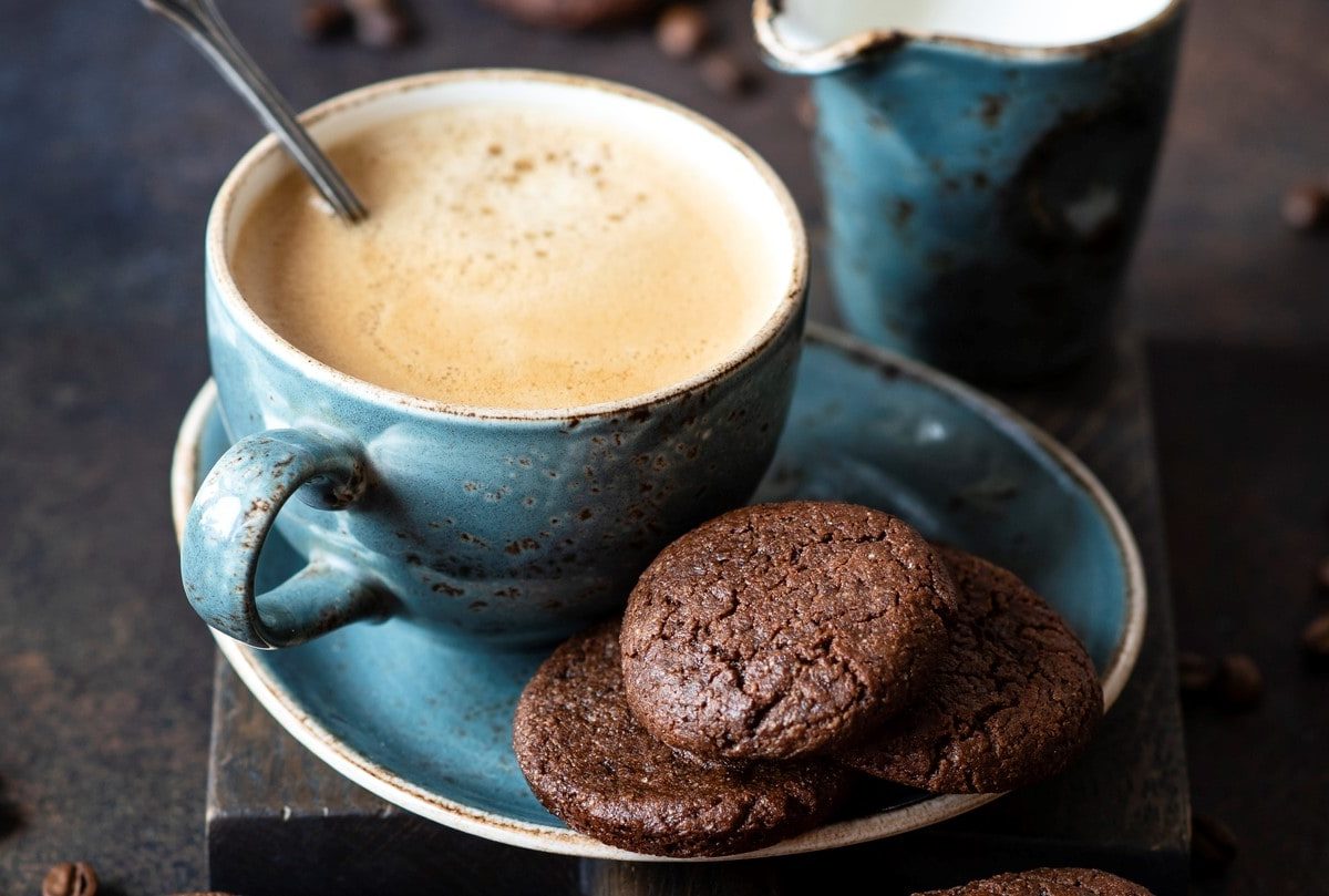 Eine gefüllte Kaffeetasse auf einer Untertasse, auf der einige vegane Schoko-Espresso-Plätzchen liegen. Drumherum verstreut sieht man Kaffeebohnen.