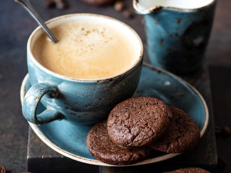 Eine gefüllte Kaffeetasse auf einer Untertasse, auf der einige vegane Schoko-Espresso-Plätzchen liegen. Drumherum verstreut sieht man Kaffeebohnen.