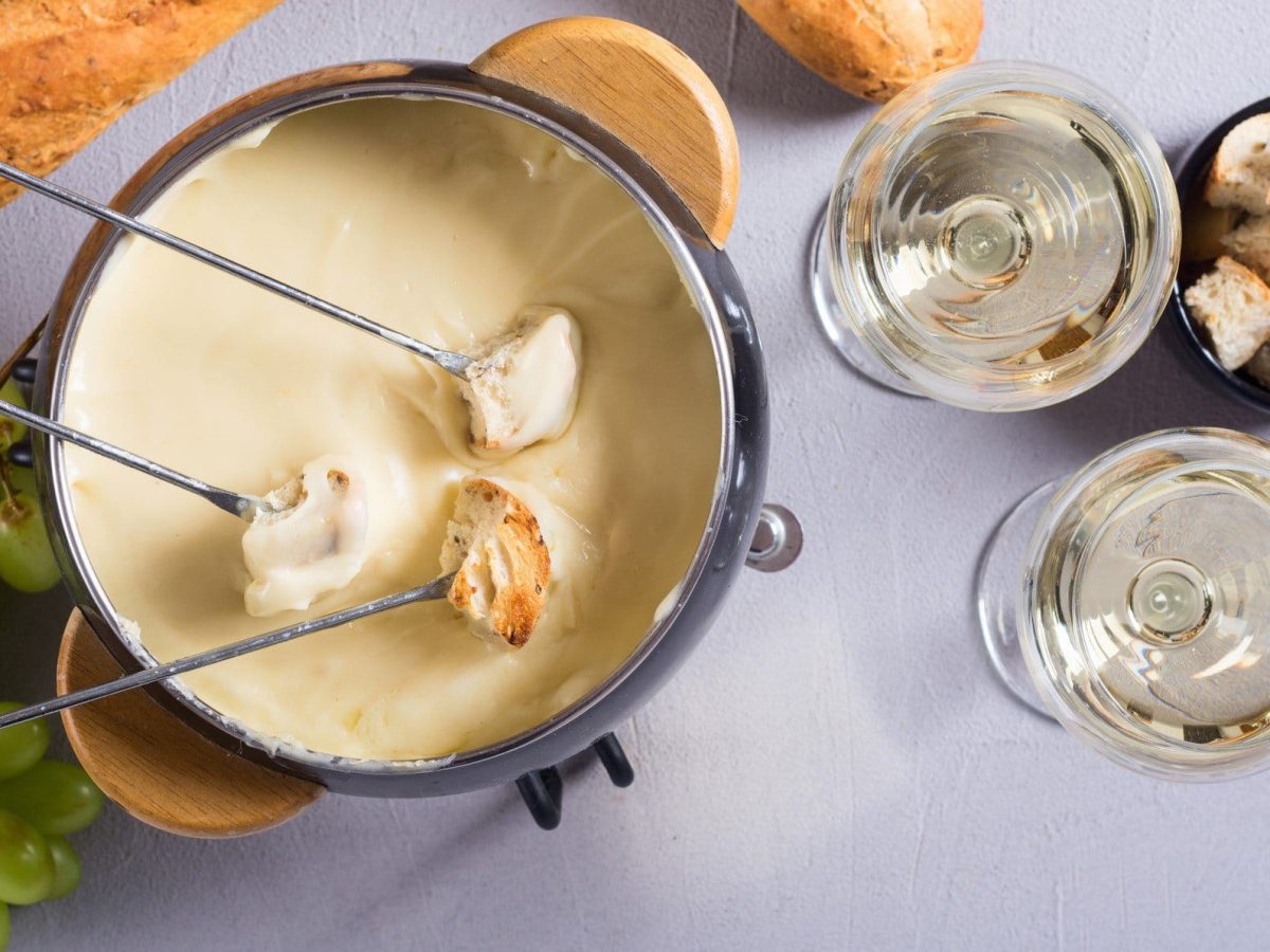 Ein Topf veganes Käsefondue mit drei Spießen, daneben zwei Gläser Weißwein, Weintrauben, Brotwürfel und ein Baguette.