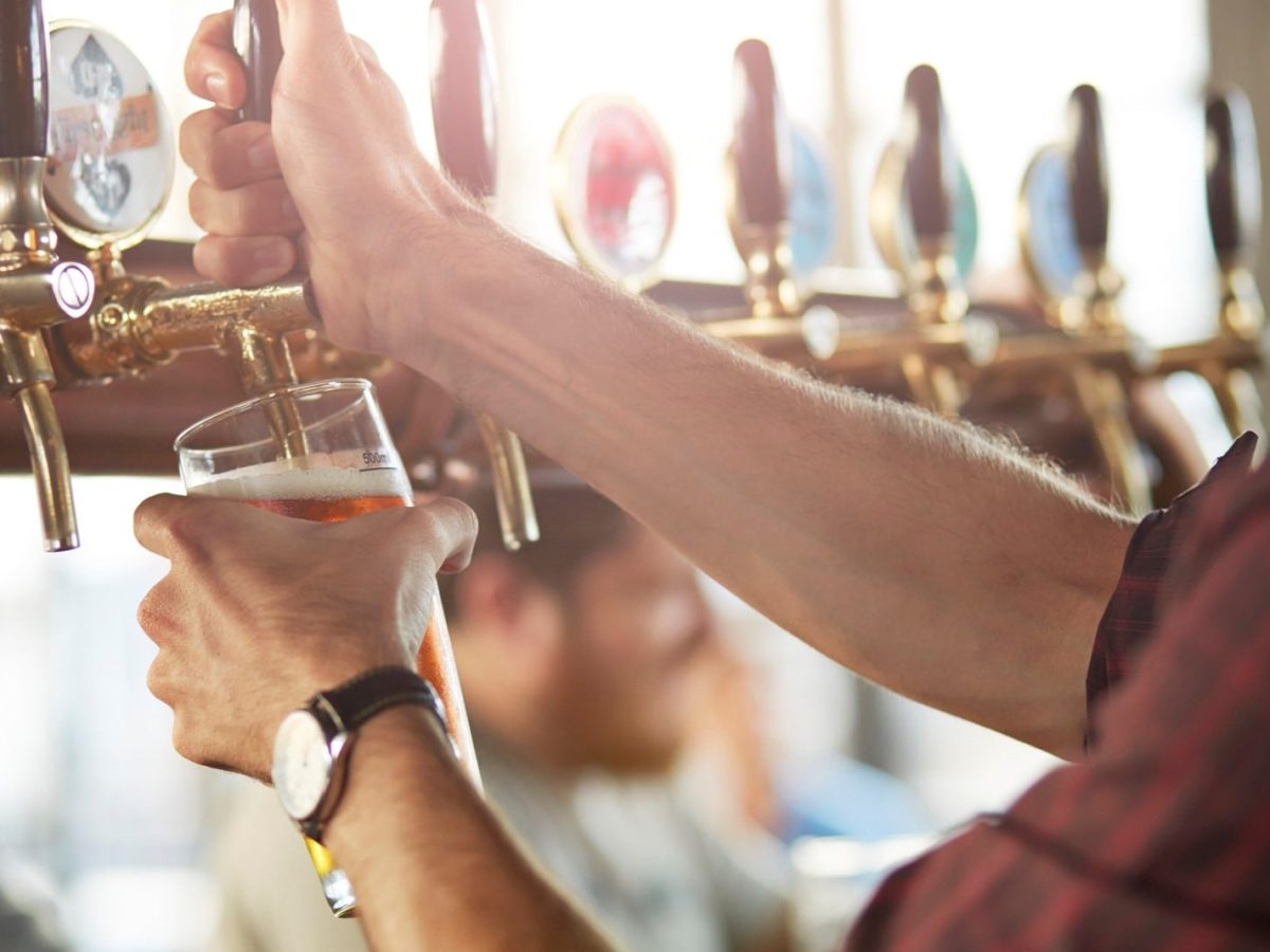 Ein Mann in einem karierten Hemd zapft helles Bier in ein großes Glas. Im Hintergrund sitzen mehrere Personen an einer Bar.