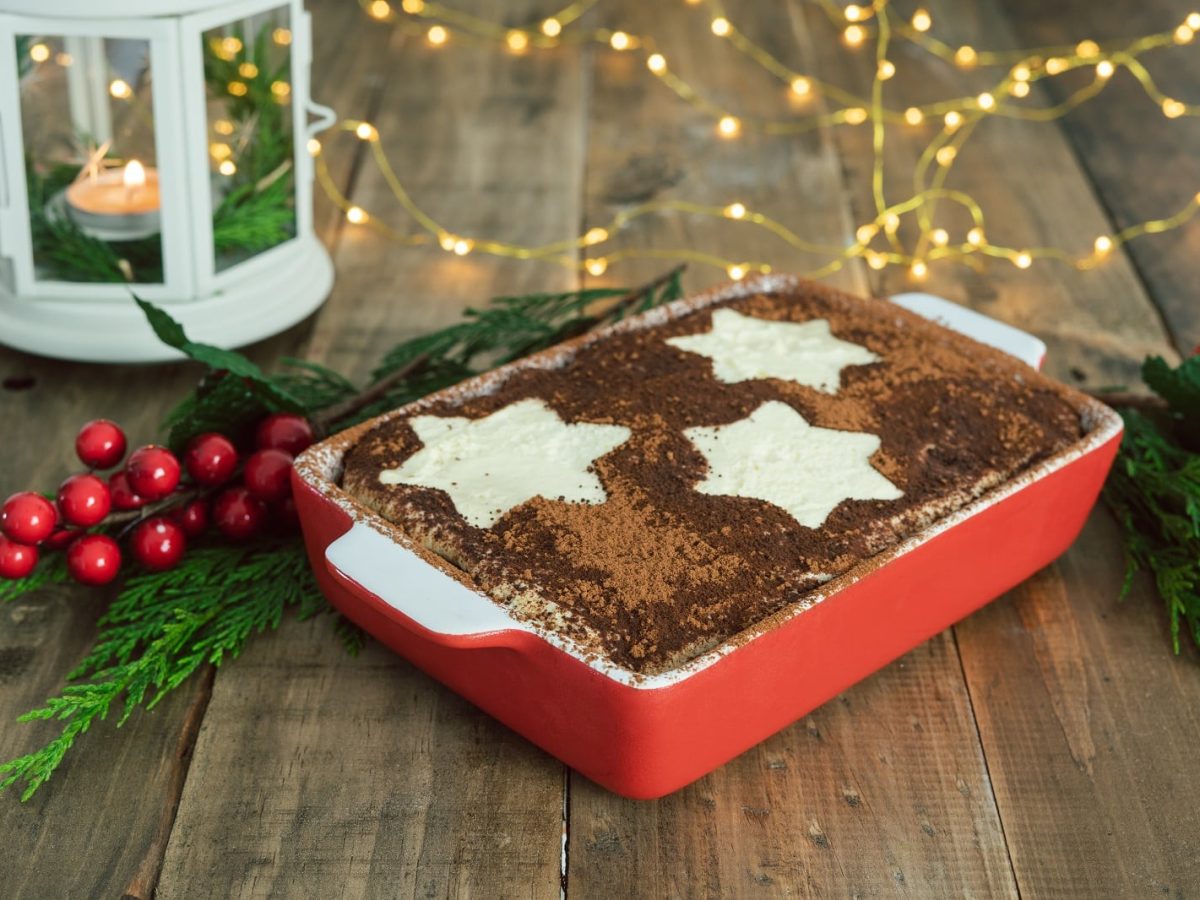 Frontalsicht: Auf einem Holutisch, der weihnachtliche geschmückt ist, steht eine große Form mit einem Tiramisu . Das ist mit Sternen aus Puderzucker dekoriert.