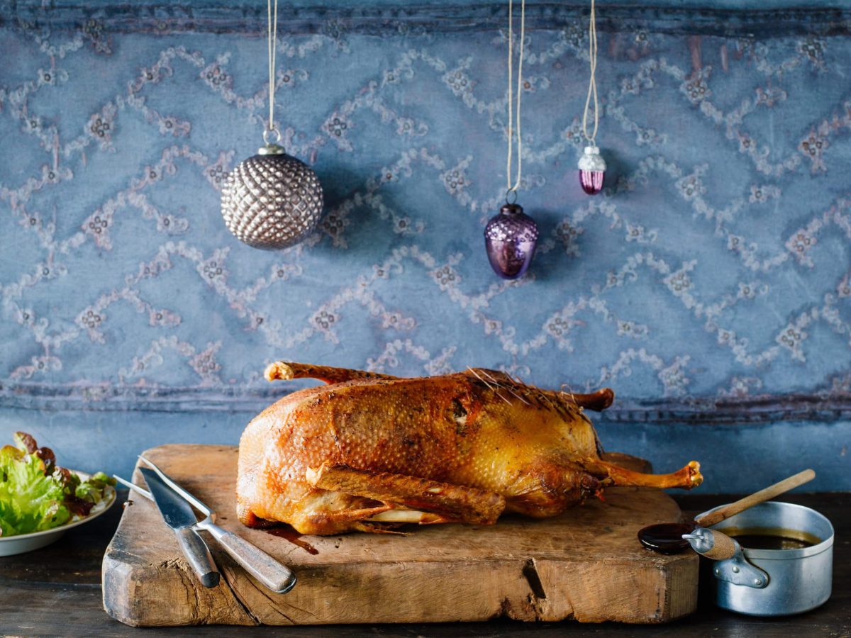 Die Weihnachtsgans liegt in der Küche auf einem dicken Holzbrett.. Im Hintergrund hängen Christbaumkugeln.