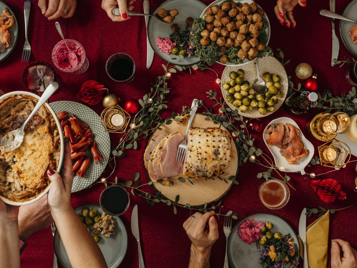 Ein festlich gedeckter Tisch mit vielen verschiedenen Speisen in der Draufsicht, dazu ein paar Hände, die sich gegenseitig Schüsseln mit Essen reichen.