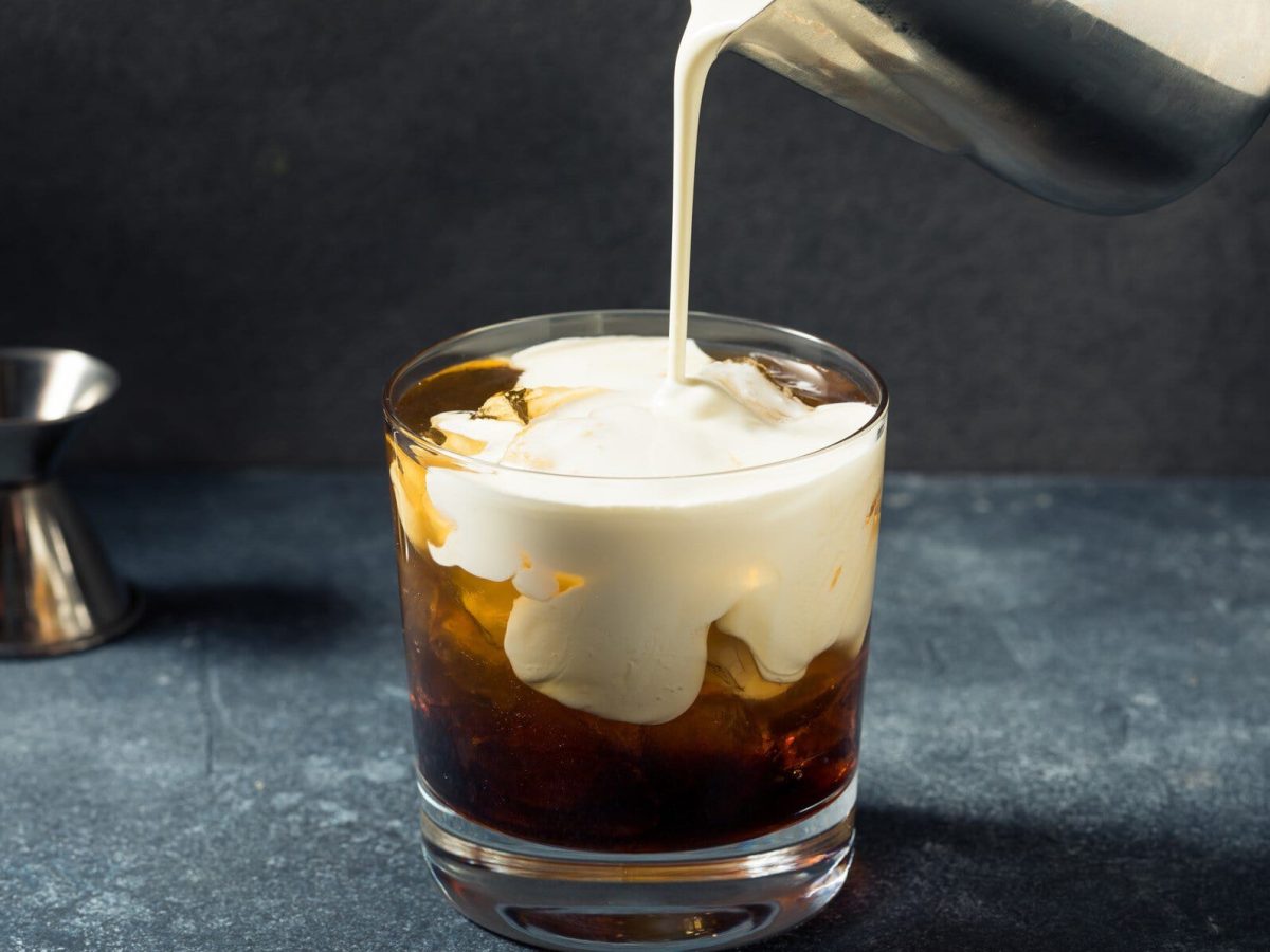 White Russian aus "The Big Lebowski" in der Entstehung im Tumbler auf Granittisch aus der Frontalansicht. Metallkännchen schüttet Sahne in das Glas. Links im Hintergrund Barmaß.