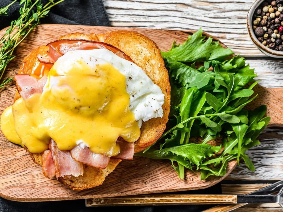 Auf einem Holzbrettchen ist ein Ei auf Bacon und Toast zu sehen. Es handelt sich um Eier Benedict