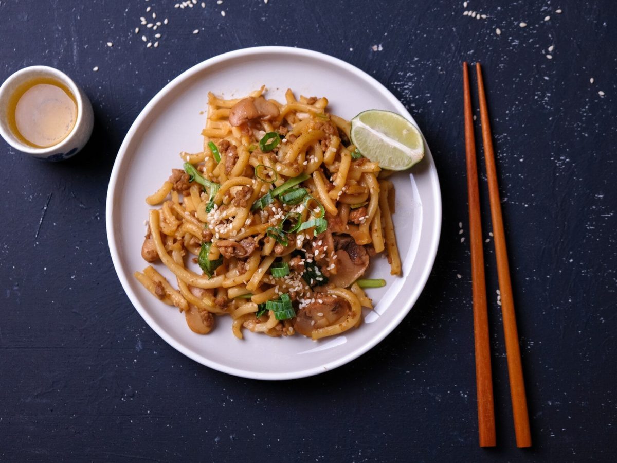 Yaki Udon mit Lauch und Sesam auf weißem Teller. Rechts Essstäbchen. Links ein Becher mit heller Flüssigkeit. Aufnahme aus der Vogelperspektive.