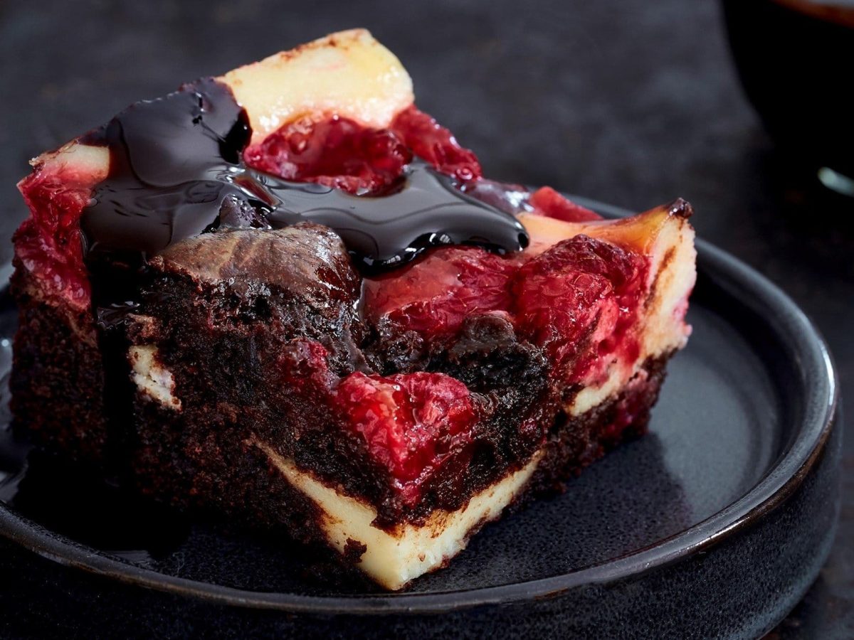 Auf einem schwarzen Porzellan-Teller liegt ein Stück der Beeren-Cheesecake-Brownies. Es ist überzogen mit Schokoladensoße.